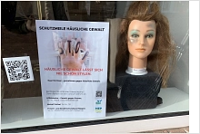 Schaufenster Stadt Coiffeur Meppen mit schlecht frisiertem Puppenkopf, der auf die Thematik Häusliche Gewalt hinweist. Slogan: "Häusliche Gewalt lässt sich nie schön stylen"